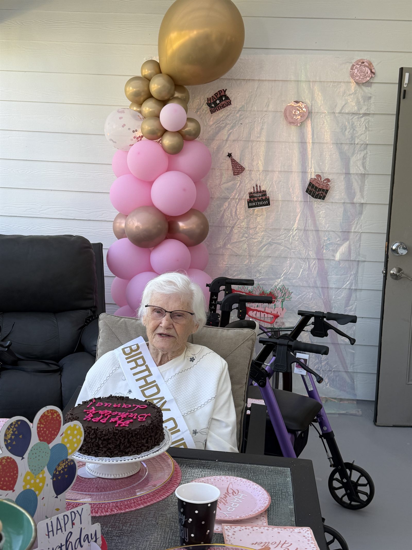 A resident in a birthday sash beside a chocolate cake and balloon arch